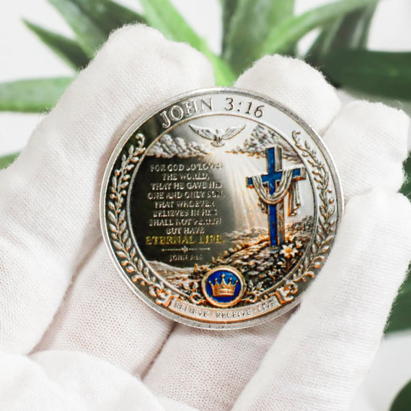 A hand holds a commemorative coin featuring John 3:16 and a cross design against a green leaf background.