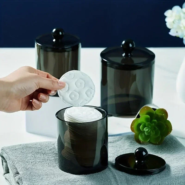 A hand is dispensing white cotton rounds from a clear glass container with a black lid onto a gray cloth.