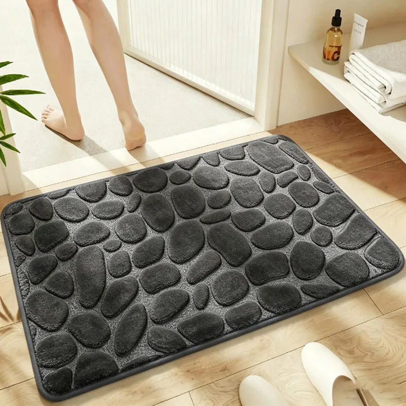 A dark gray, pebble-textured bath mat is placed on a wooden floor next to a white door frame.