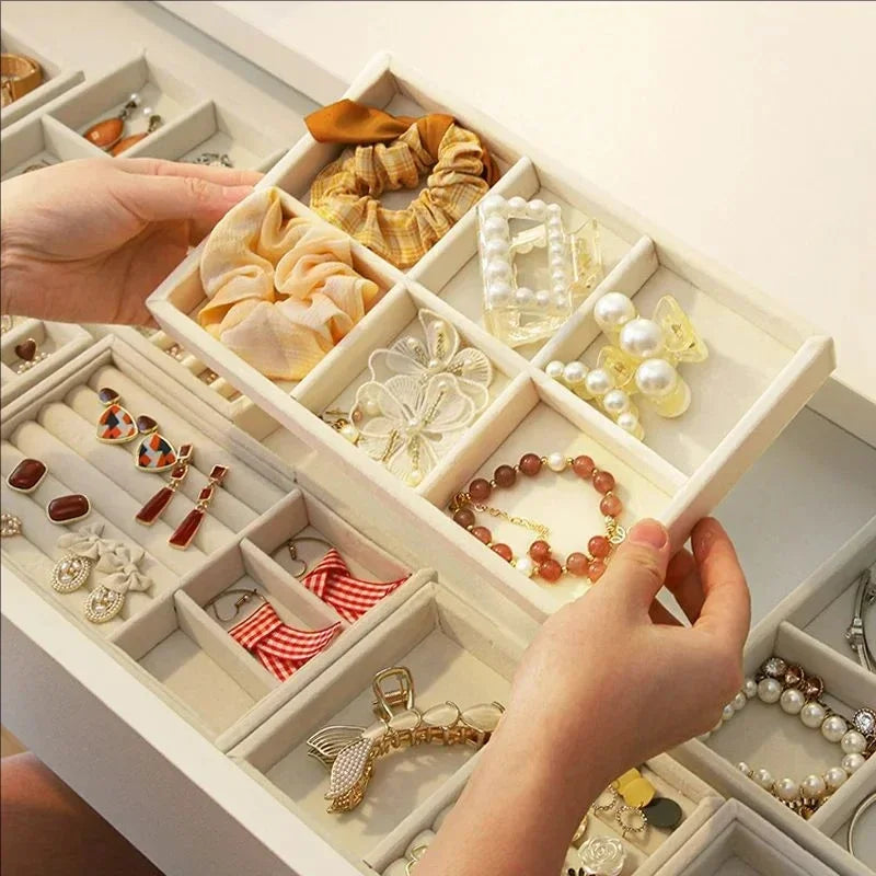 A hand is organizing various pieces of jewelry, including rings, earrings, and bracelets, in a white velvet tray with soft li