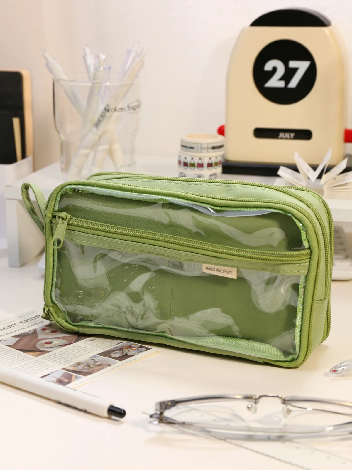 A light green transparent pencil pouch resting on a white surface next to various stationery items.