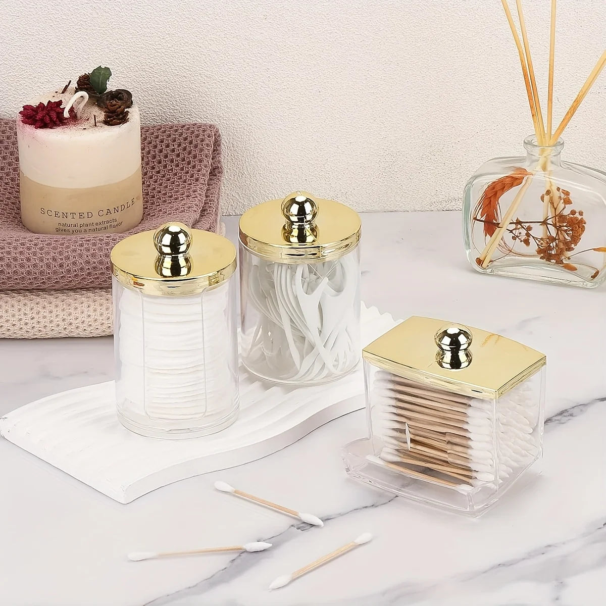 A clear apothecary jar dispenser with a golden lid holds white cotton balls and swabs on a marble surface.