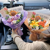 A handmade crochet flower bouquet with various textured flowers in shades of orange, purple, and yellow is displayed in an op