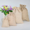 Mehrere natürliche Jute- und Leinen-Geschenktüten verschiedener Größen neben einem Topf mit lila und gelben Blumen