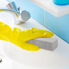 A yellow rubber glove is holding a gray pumice stone near a faucet on a white sink.