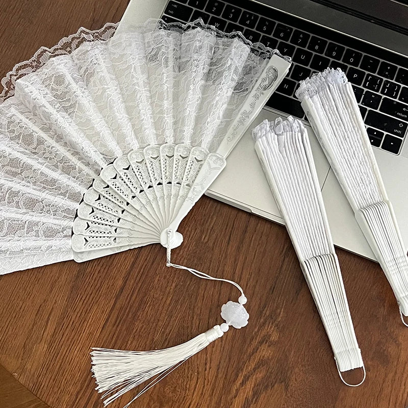 White lace fabric folding hand fan with delicate pleats and a tassel resting on a wooden surface.