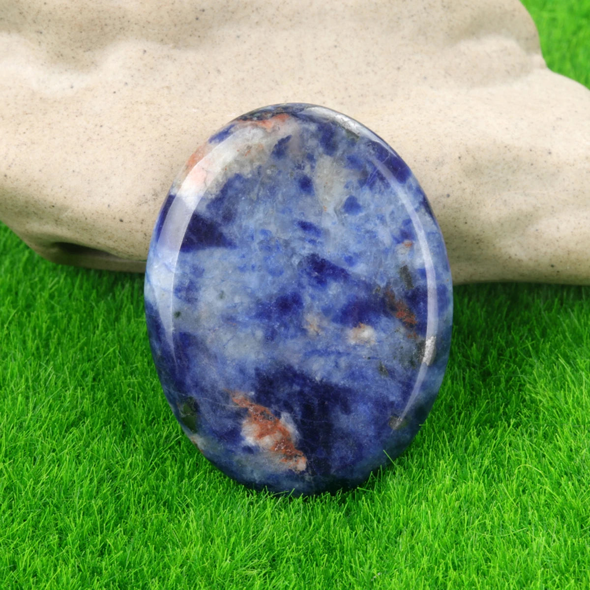 A smooth, oval blue and purple quartz stone rests on green grass next to a hand.