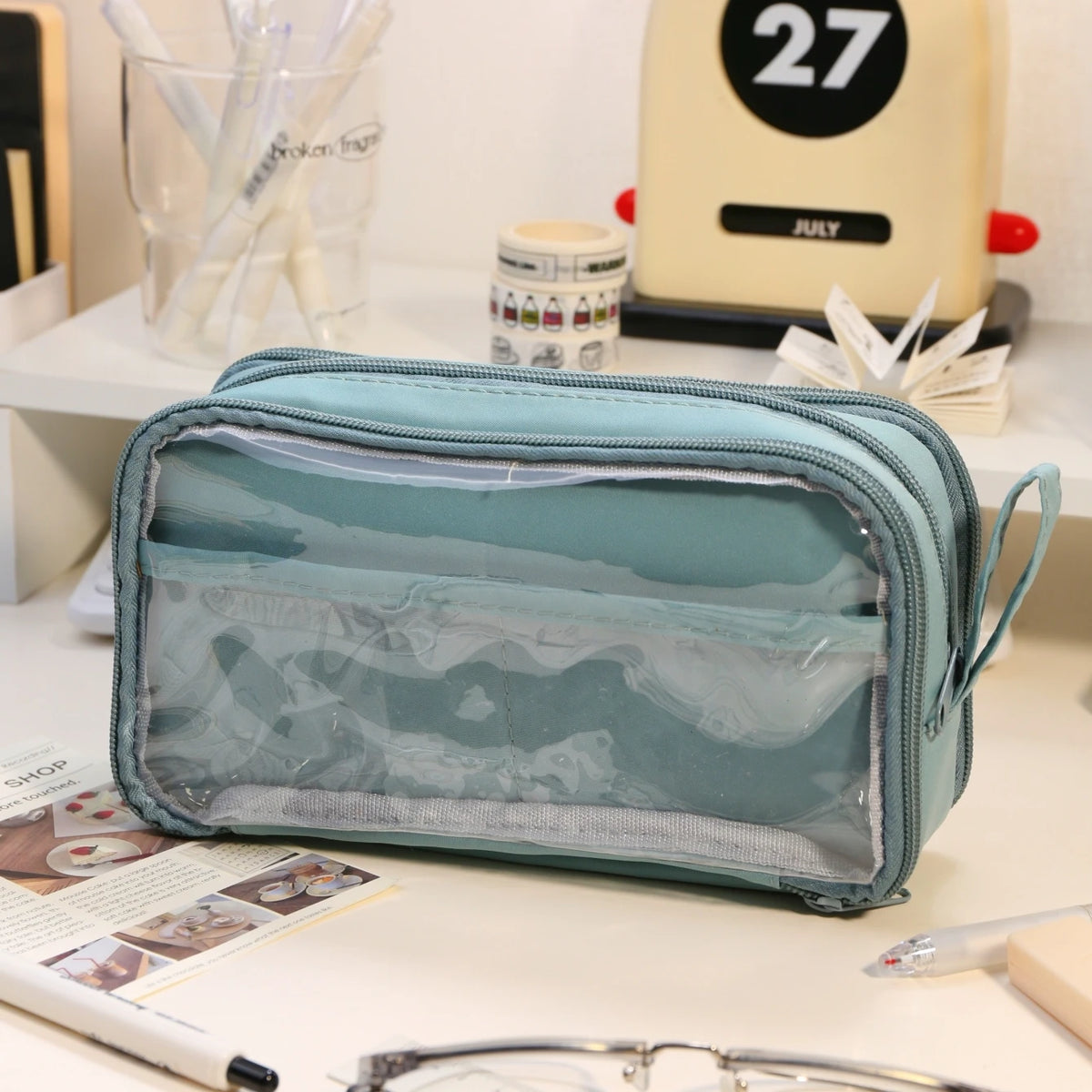 A light blue transparent pencil pouch is displayed on a white surface next to other stationery items.