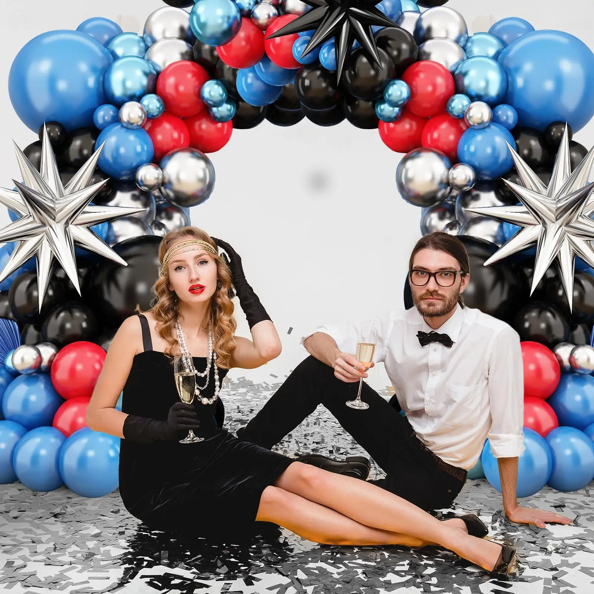 A woman in a black dress and gloves lies on the ground next to a man, surrounded by a balloon garland of blue, red, and black