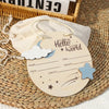 Etiqueta de madera con forma de luna creciente y estrellas, con el texto Hello World y campos para nombre, fecha y peso