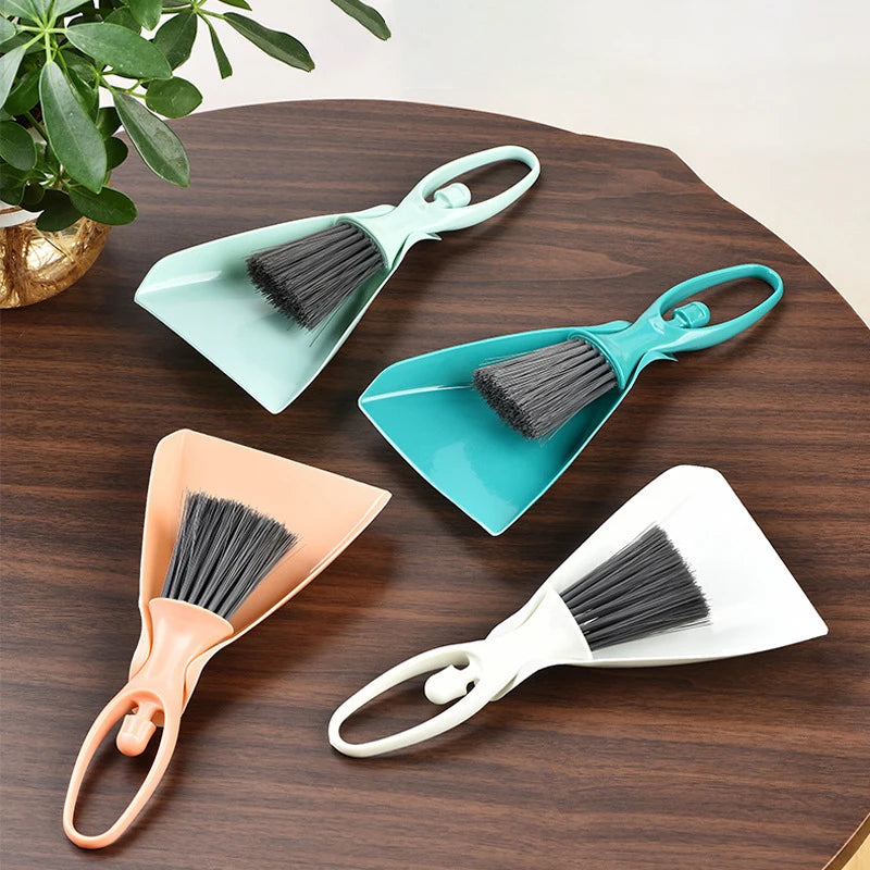 A set of three mini brooms and dustpans in teal, orange, and white resting on a dark wooden surface.