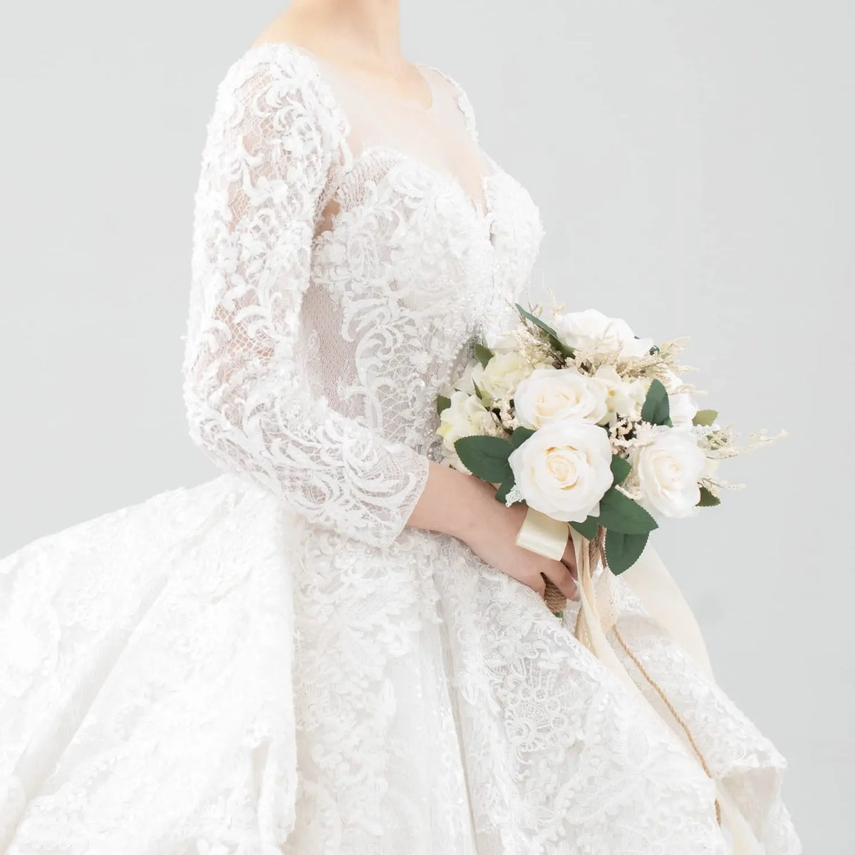 A bride wearing a white lace wedding dress holds an elegant bouquet of white roses and greenery.