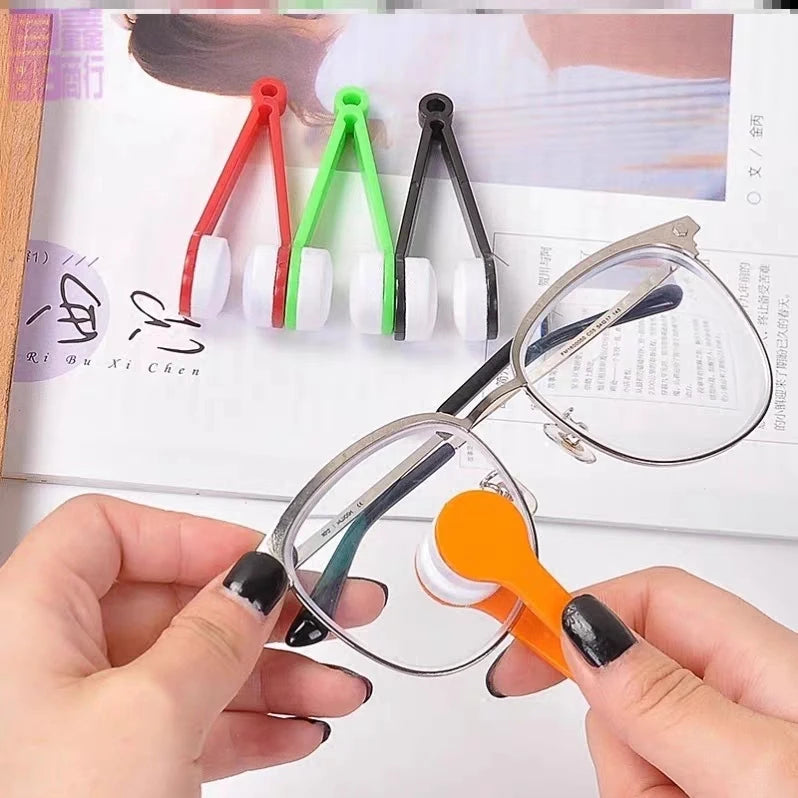 A hand holds a pair of eyeglasses with an orange cleaning brush and white bristles, next to a set of green and red microfiber
