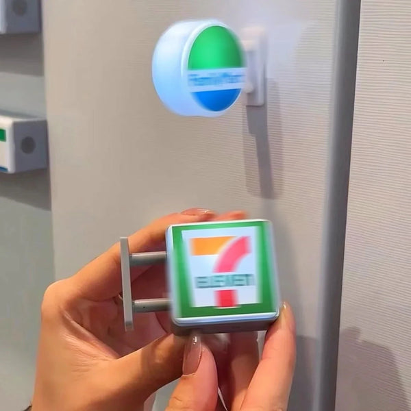 Une main tient un aimant réfrigérateur miniature vert et blanc décor contre une surface gris clair.