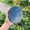A hand holds a round black obsidian scrying mirror reflecting the sky and surrounding greenery.