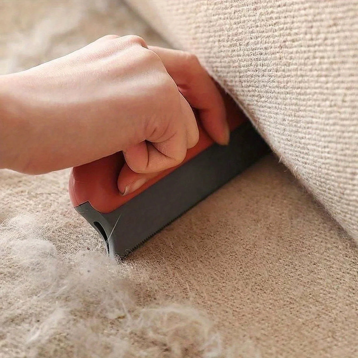 A hand is using a gray pet hair remover brush on a beige carpet, removing pet hair.