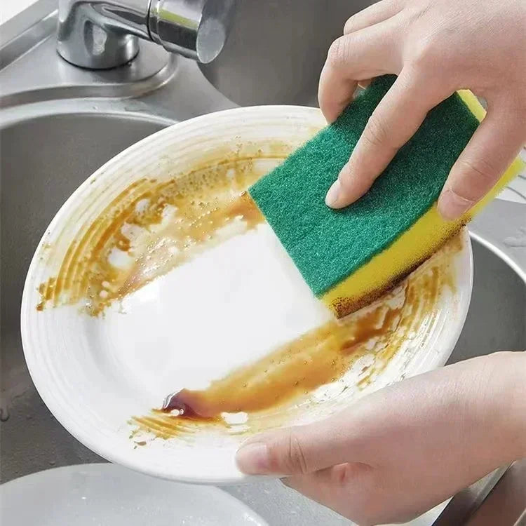 A hand is using a green and yellow double-sided magic cleaning sponge to wash food residue from a white plate in a kitchen si