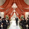 A wedding ceremony setup featuring a bride and groom standing on an aisle lined with white flowers and candles under draped o