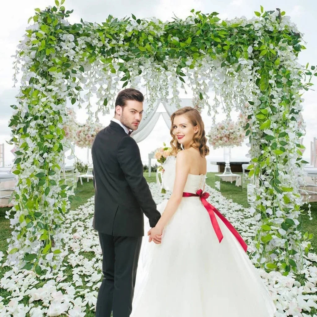 A bride and groom stand under a lush green and white floral archway for a wedding photo.