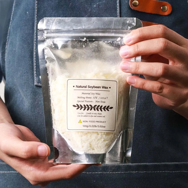 A hand holds a clear plastic pouch containing white natural soybean wax flakes for candle making.
