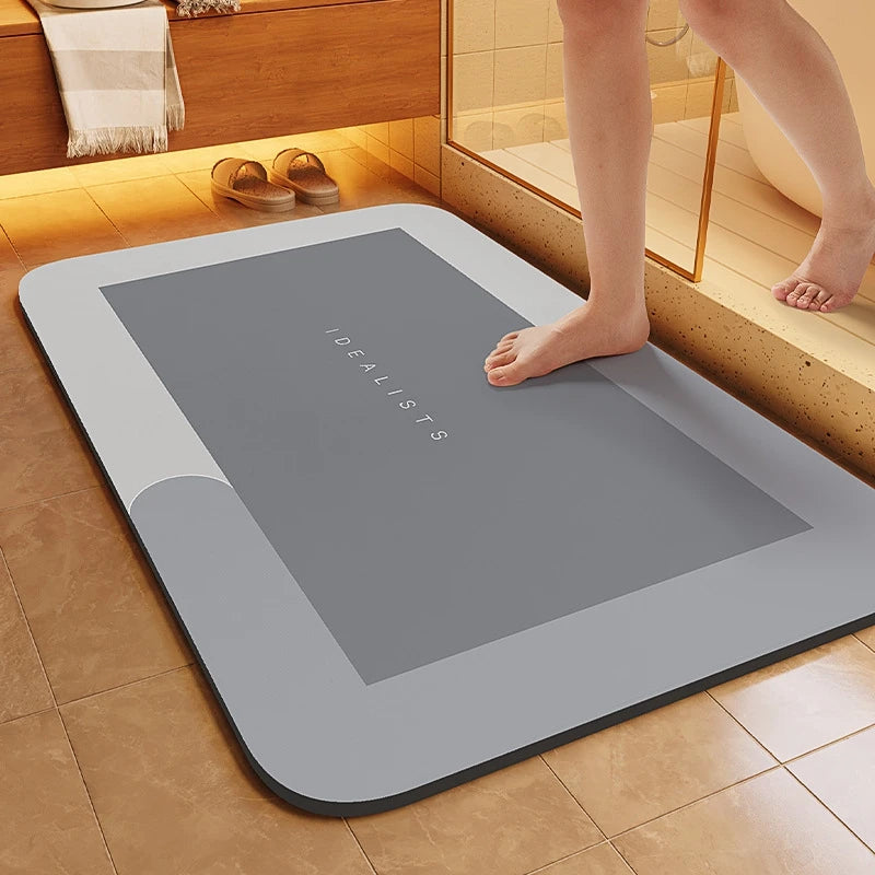 A gray non-slip bath mat with the word IDEALISTS printed on it is placed on a tiled floor next to a wooden cabinet.