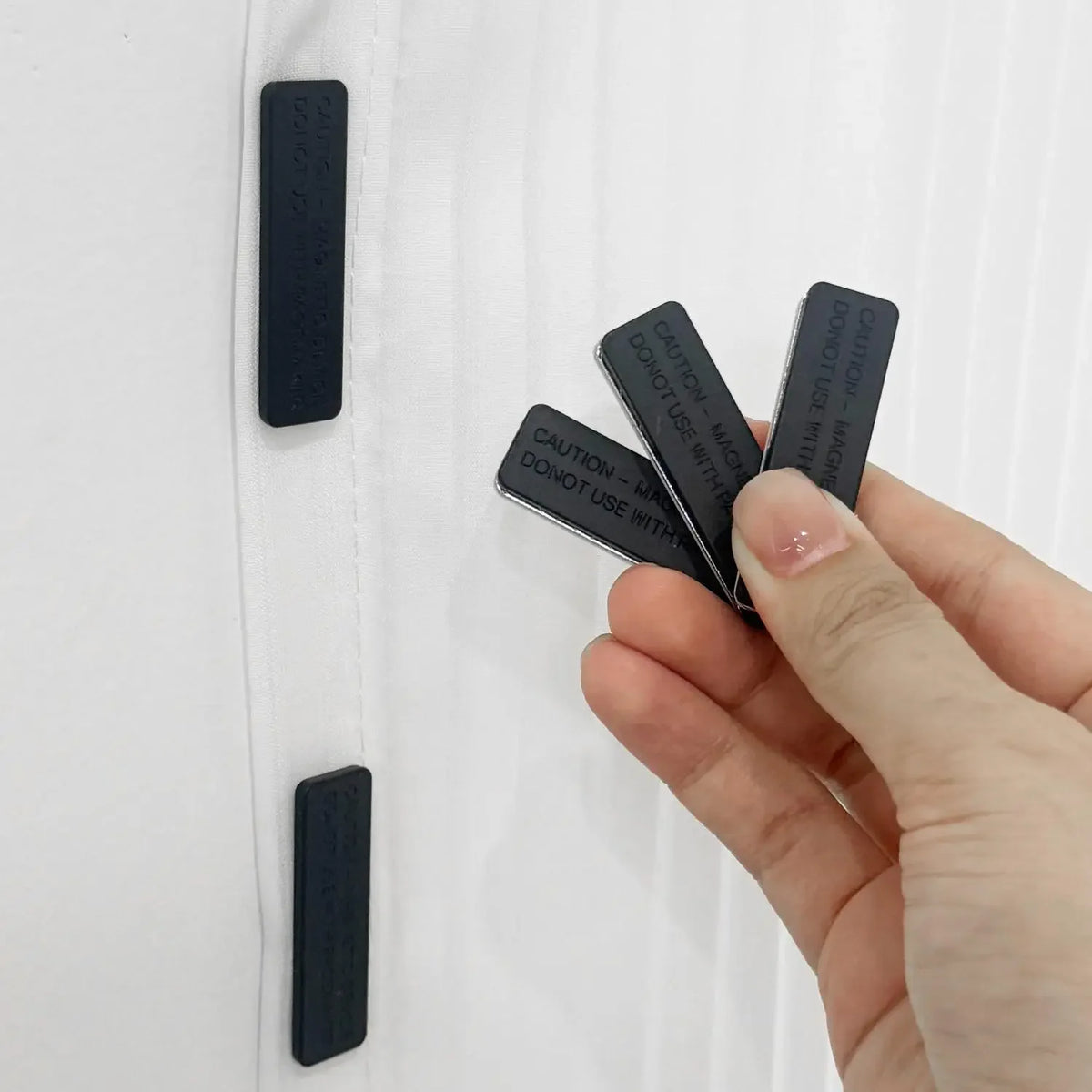 A hand is holding a black magnetic curtain clip with a warning label, demonstrating its use.