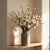 A vase filled with artificial white plum blossoms displayed on a wooden shelf.