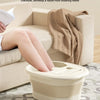 A beige portable foldable foot bath tub is shown next to a sofa, with a person's legs soaking in the bath.