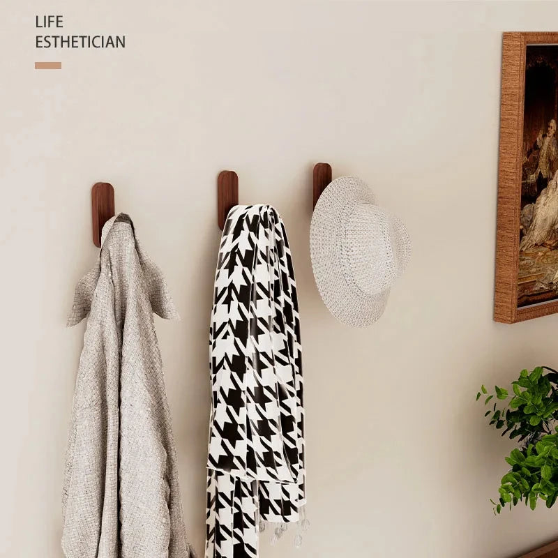 Three walnut wood wall hooks display a gray towel, a black and white patterned scarf, and a light gray straw hat.