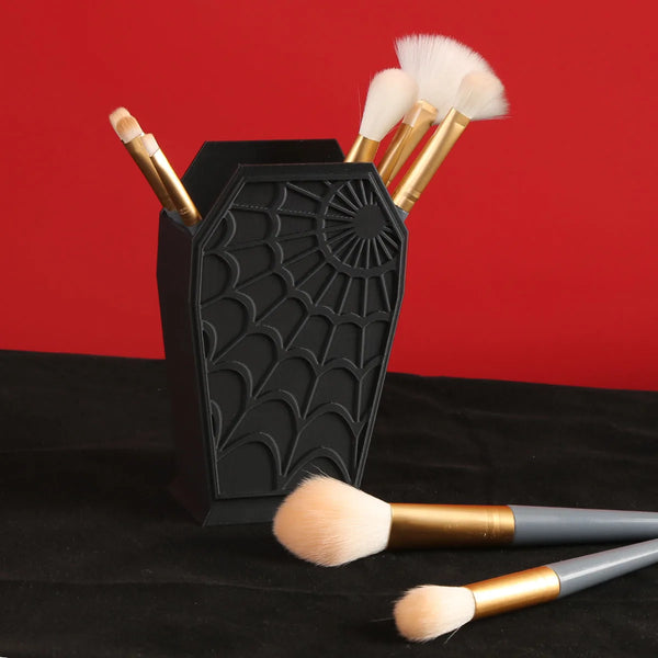 Black coffin-shaped makeup brush holder with a spider web design and several gold-handled makeup brushes.
