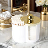 A white plastic bathroom organizer holds cotton swabs and makeup accessories with a gold lid on a marble surface.