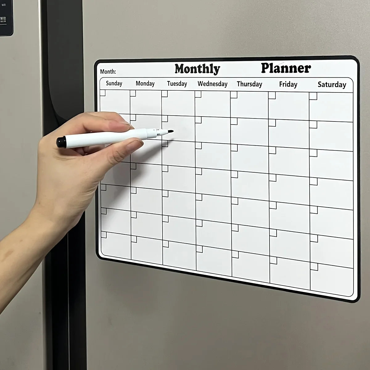 Eine Hand schreibt in einem monatlichen Planerkalender, der auf dem Kühlschrank angezeigt wird