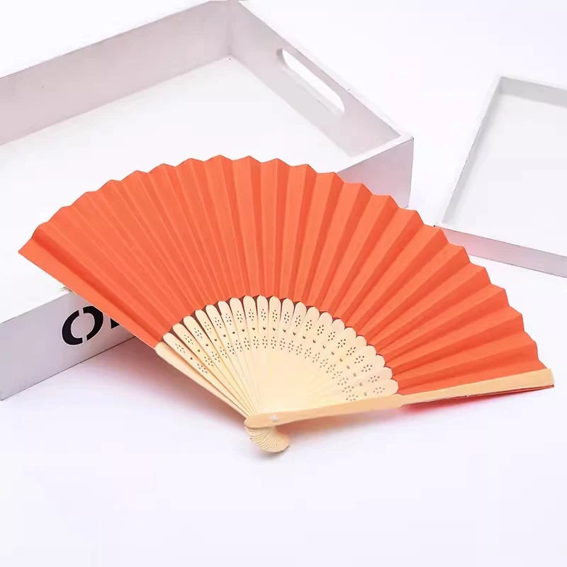 Ventilateur pliable à main orange en bambou et papier avec un cadre en bois clair.