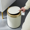 A hand is lifting the lid of a white and gold durable press-type trash can.