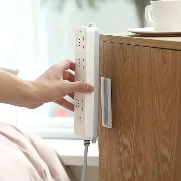 A hand is placing a white desktop cable organizer holder onto a wooden cabinet, showing the self-adhesive mechanism.