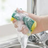 A hand is holding a green and yellow double-sided scouring sponge under running water in a kitchen sink.