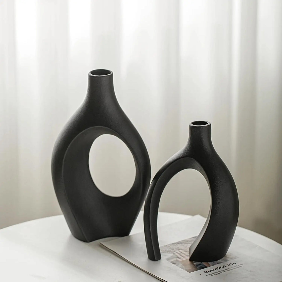 A black modern minimalist ceramic vase with an elongated, curved shape is displayed on a white surface.