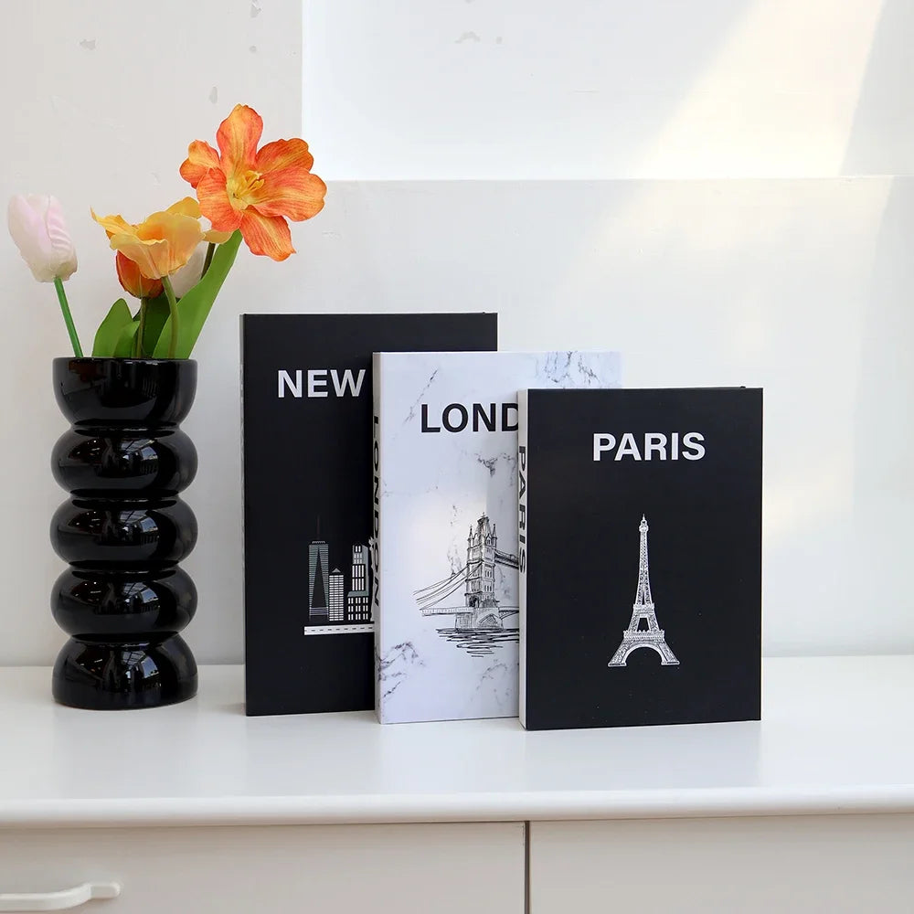 Three decorative fake books featuring city skylines, including New York, London, and Paris, displayed on a white shelf.