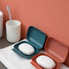 A white portable travel soap box with an open lid rests on a pink wall next to a white cup and a white towel.