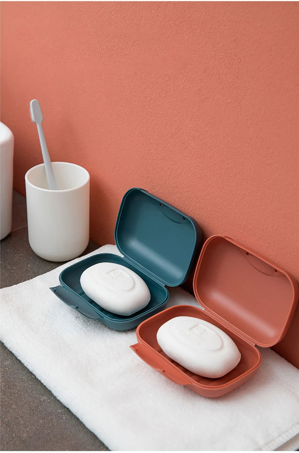A white portable travel soap box with an open lid rests on a pink wall next to a white cup and a white towel.