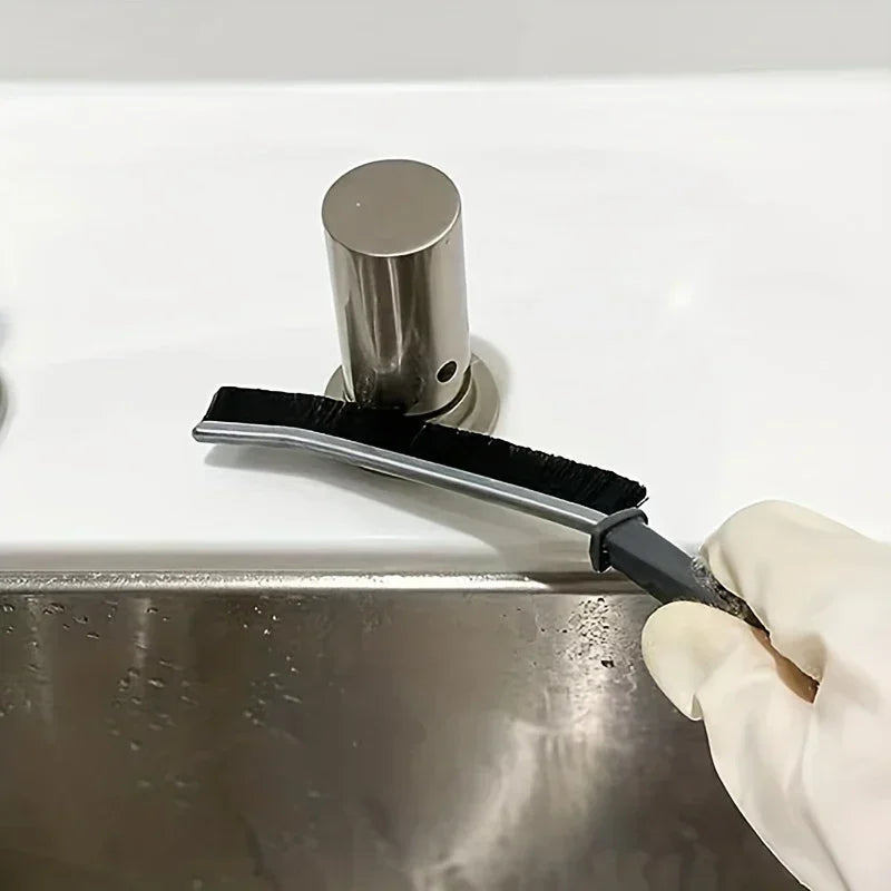 A hand is using a black hard-bristle crevice cleaning brush to clean a stainless steel sink.