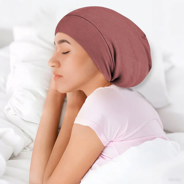 A woman wearing a dusty rose adjustable silk-lined night cap rests on white bedding.