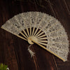 A delicate white lace folding hand fan with a wooden handle and pleated paper blades resting on a dark wooden surface.
