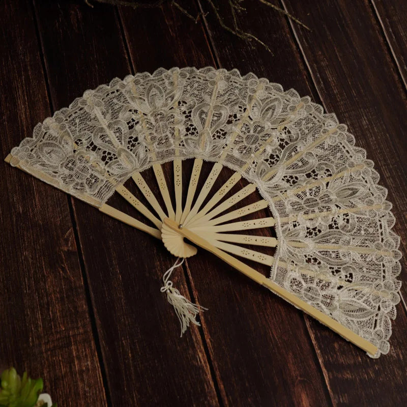 A delicate white lace folding hand fan with a wooden handle and pleated paper blades resting on a dark wooden surface.