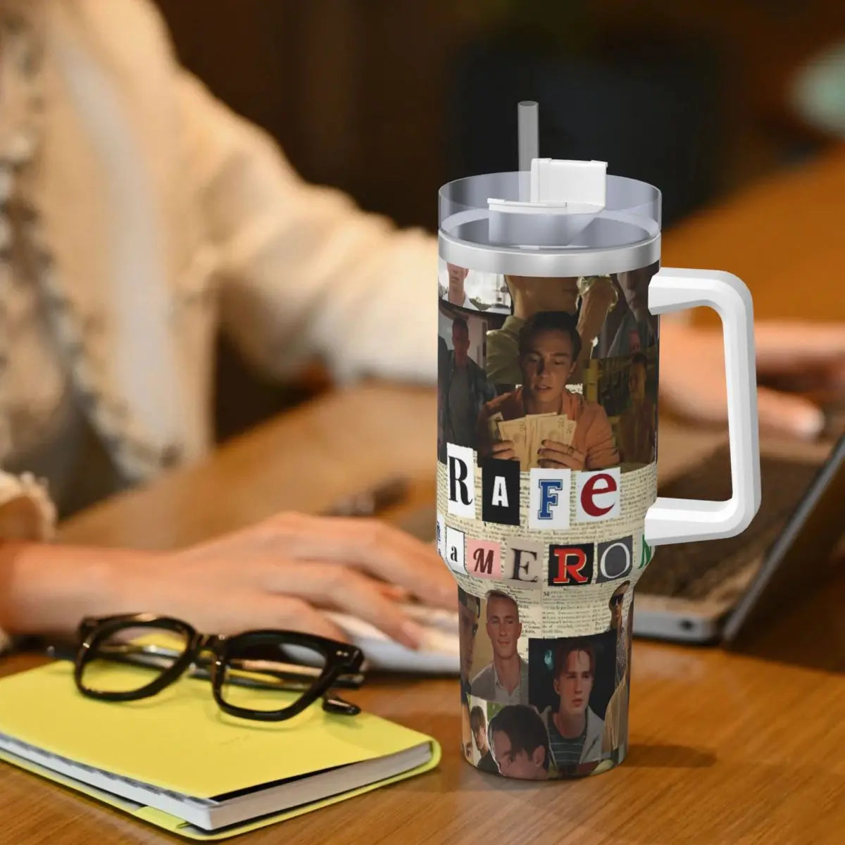 A stainless steel travel tumbler mug featuring a collage of faces and text on a wooden table.