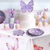 Un gâteau d'anniversaire rose décoré de toppers papillons violets sur une nappe blanche.
