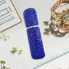 A tube of sparkling blue diamond glass perfume spray bottles resting on a white surface next to some dried flowers.