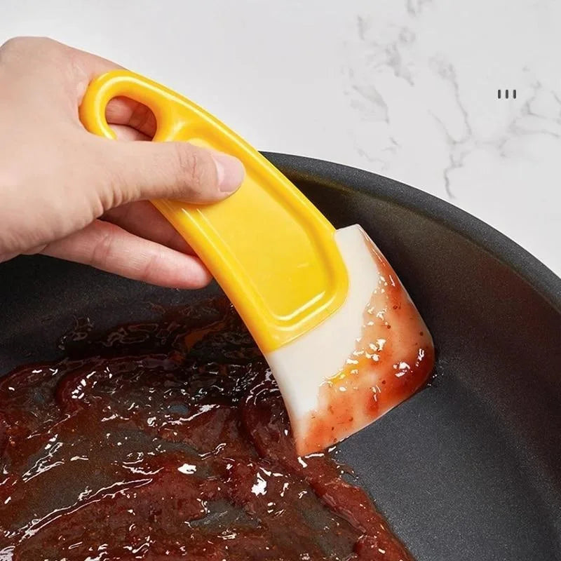 A hand is using a yellow silicone kitchen scraper to move reddish-orange food from a dark pan.