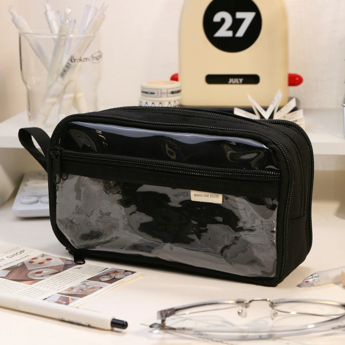 A black transparent pencil pouch is displayed on a white surface next to various writing tools and accessories.