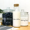 Three glass jars and a bottle containing stain remover fabric softener and washing soda on a wooden surface